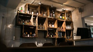 Angolo bar con mensole in legno piene di bottiglie e bicchieri di diverse tipologie.