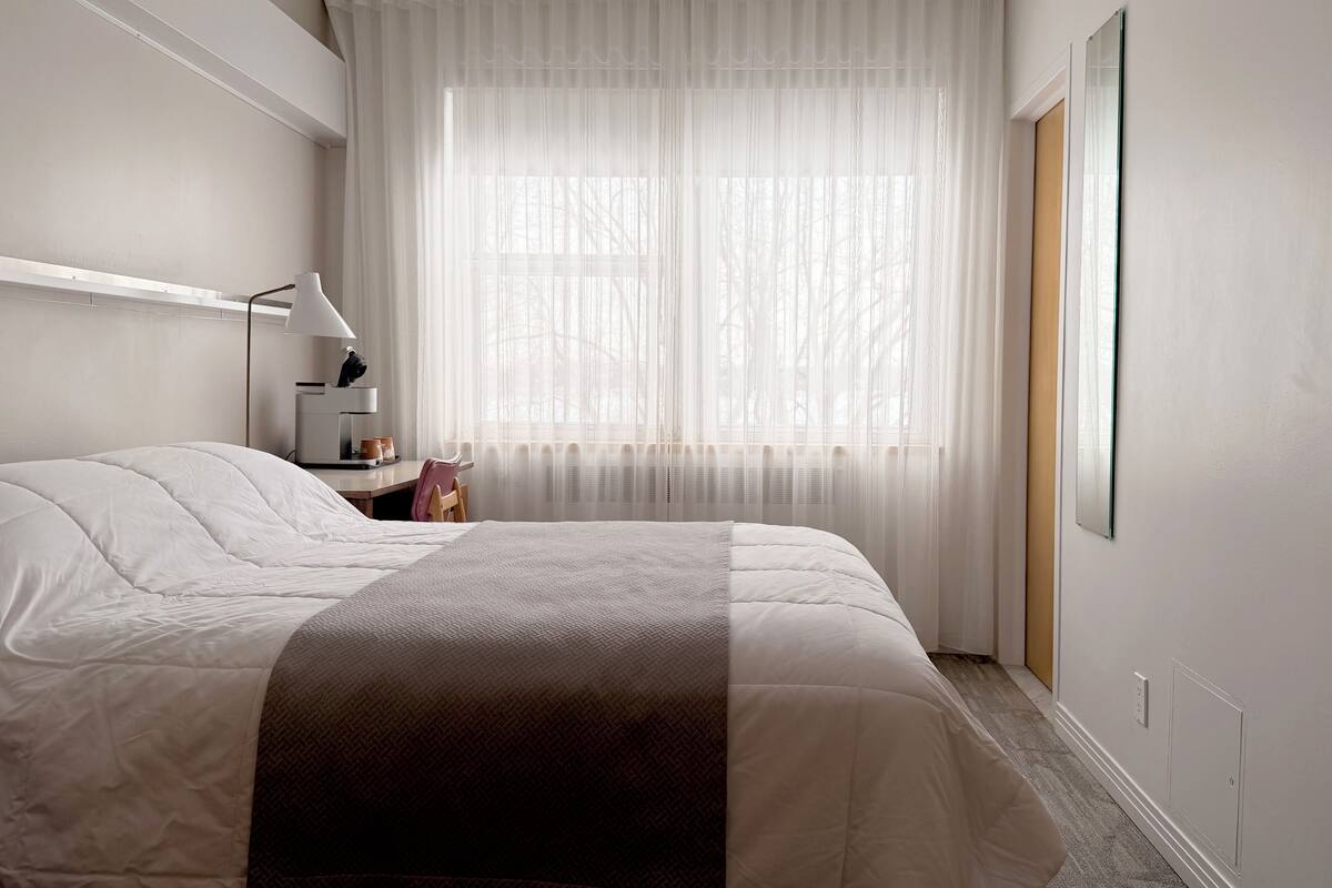 Habitación de hotel con cama, escritorio con lámpara, una silla y un ventanal con cortinas traslúcidas.