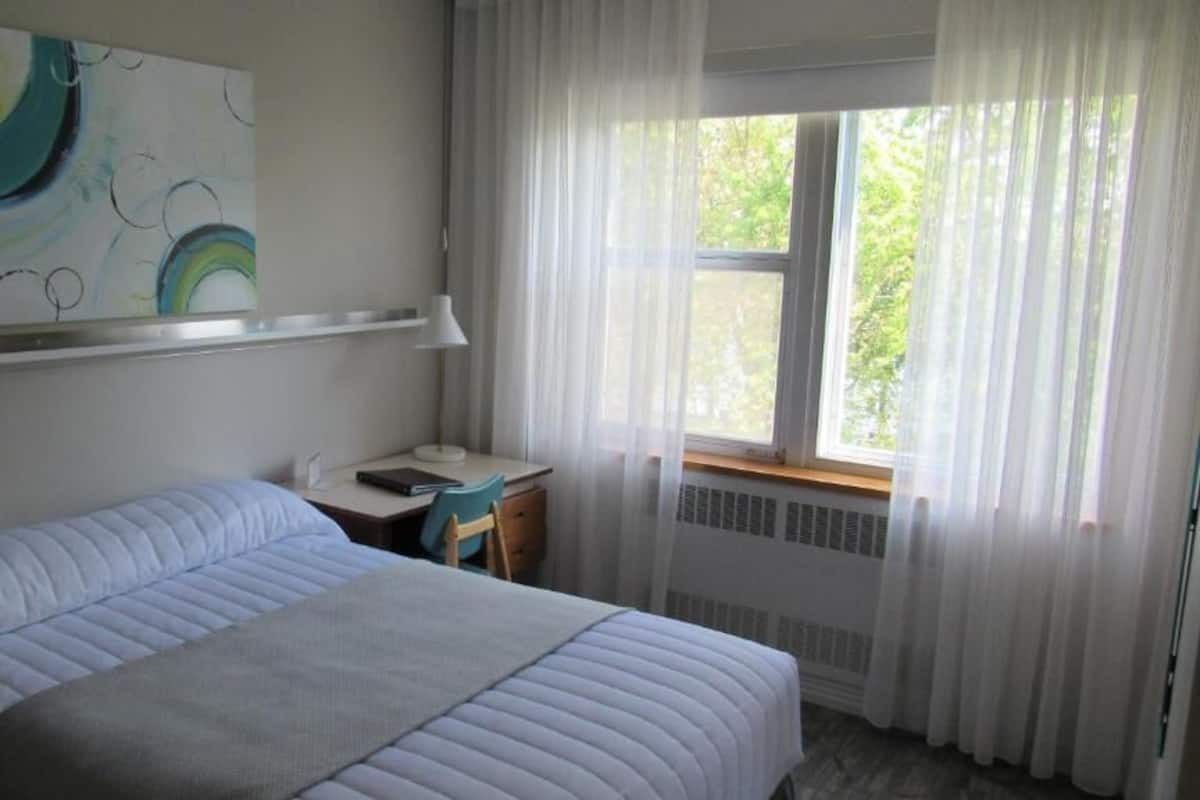 Un dormitorio con cama, escritorio, silla, ventana con cortinas y un cuadro en la pared.