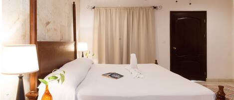 A hotel room with a bed, a nightstand with a lamp, a vase with a plant, and a door.