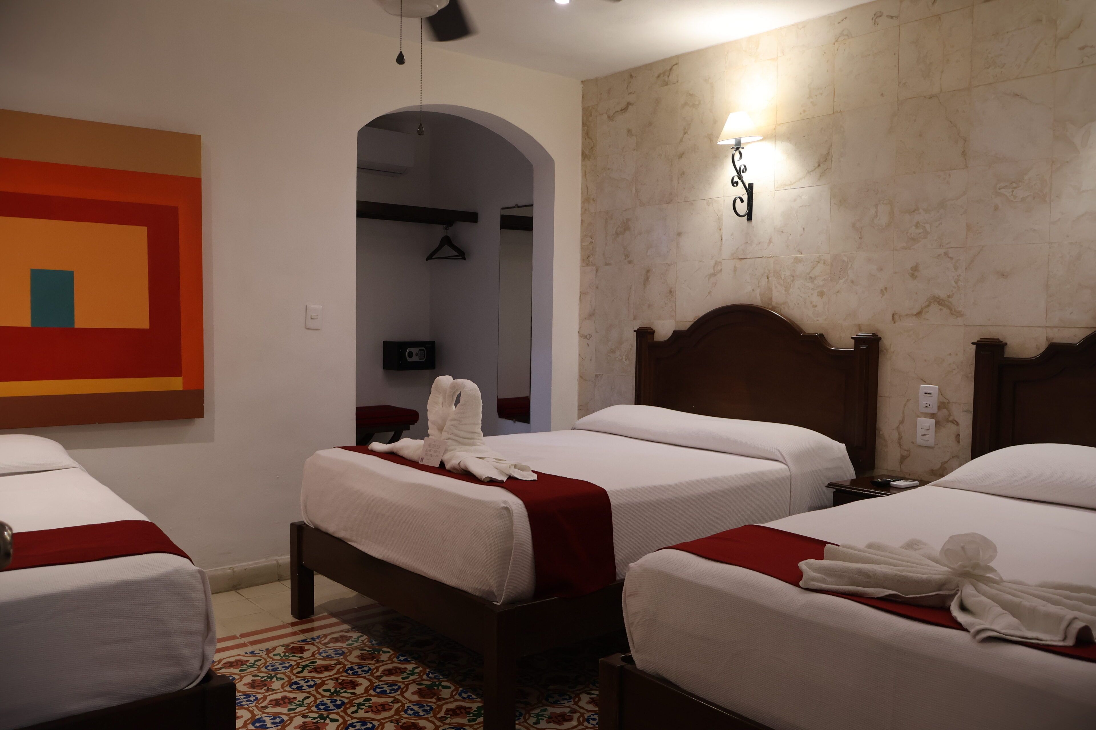 Une chambre d’hôtel avec trois lits, un tableau abstrait aux couleurs rouge et orange, un mur en marbre et un luminaire suspendu.