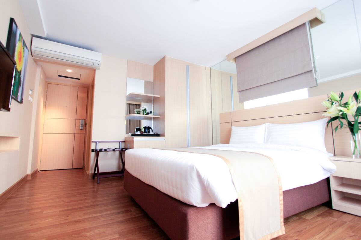 A hotel room with a bed, bedside table, and a small desk with a chair. There is a window with a blind, a wall-mounted air conditioner, and a mirror above the bed.