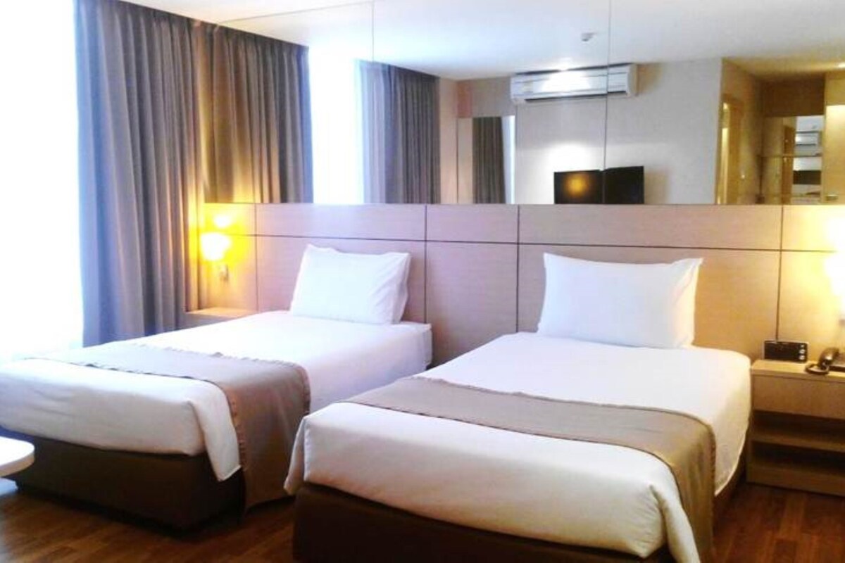 A hotel room with two beds, a bedside table with a lamp, an air conditioning unit, and a large mirror.