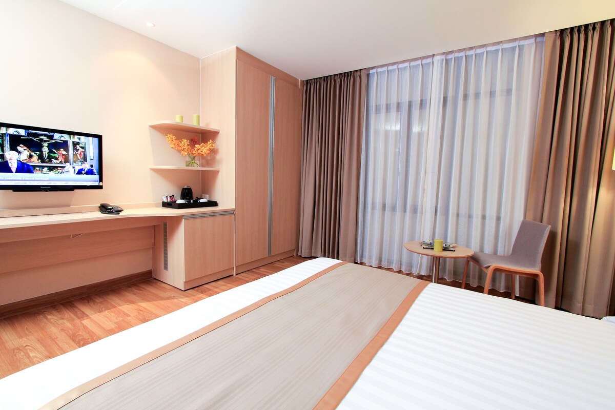 A hotel room with a bed, a TV mounted on the wall, a desk with a chair, and a small table with a chair.
