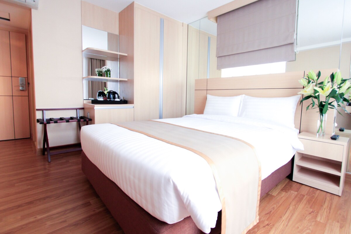 A modern hotel room with a large bed, wooden flooring, built-in wardrobe, bedside table, and a small desk with a chair.