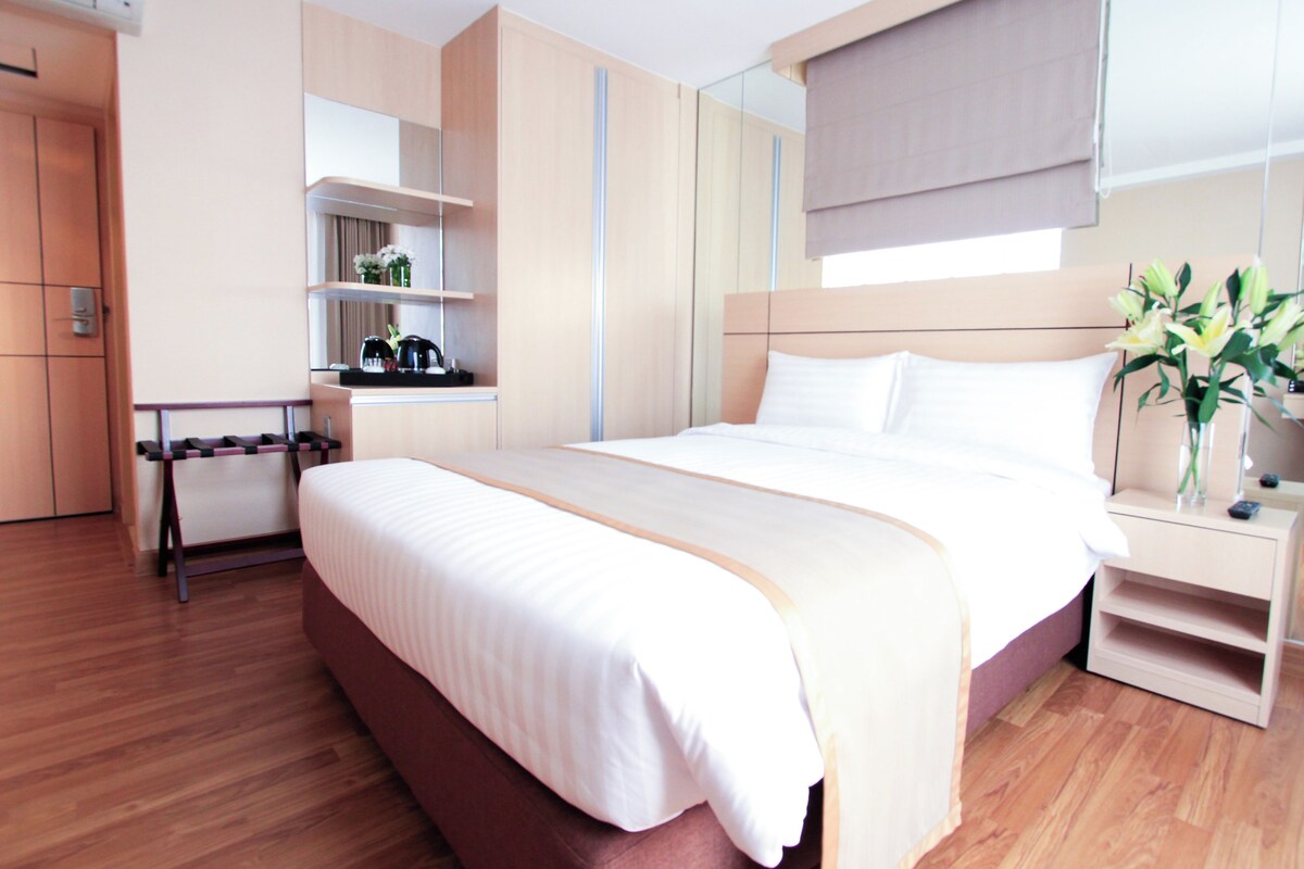 A modern hotel room with a large bed, wooden flooring, a built-in desk, and a bedside table with a vase of flowers.