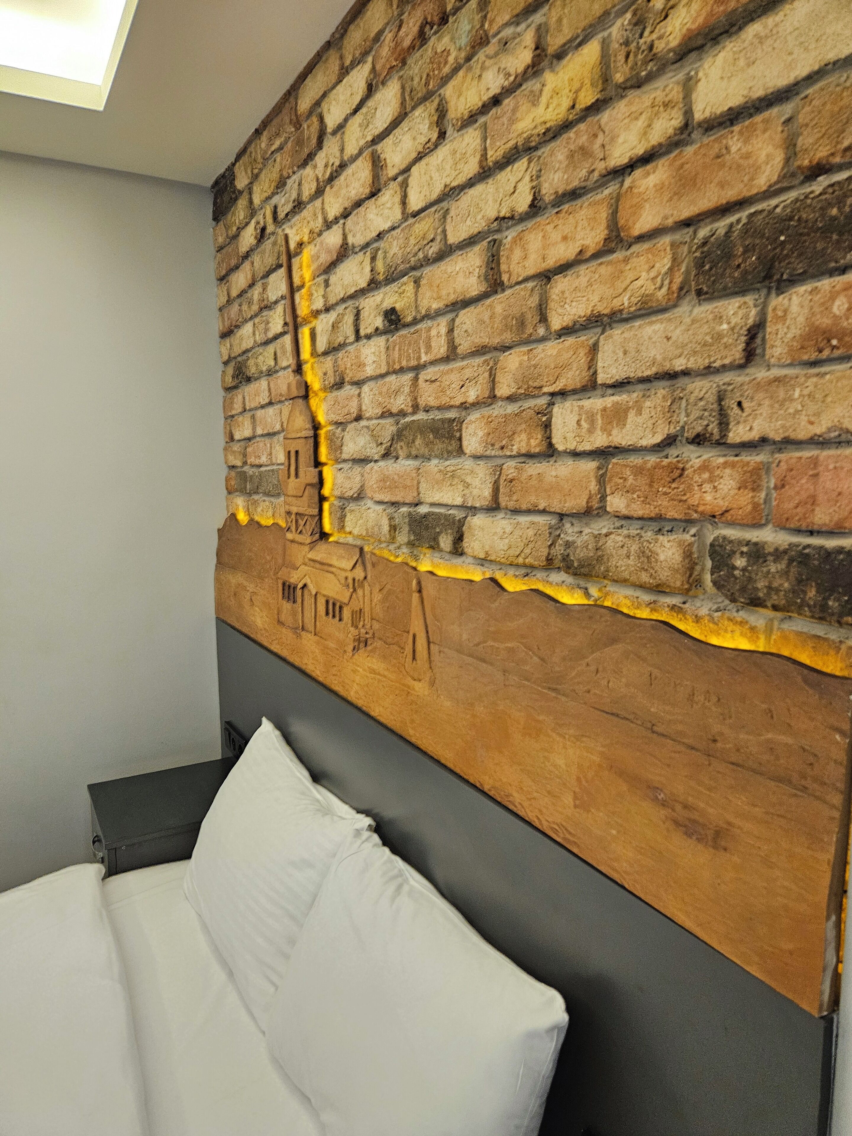 A room with a brick wall, a bed with white bedding, and a nightstand with a lamp.