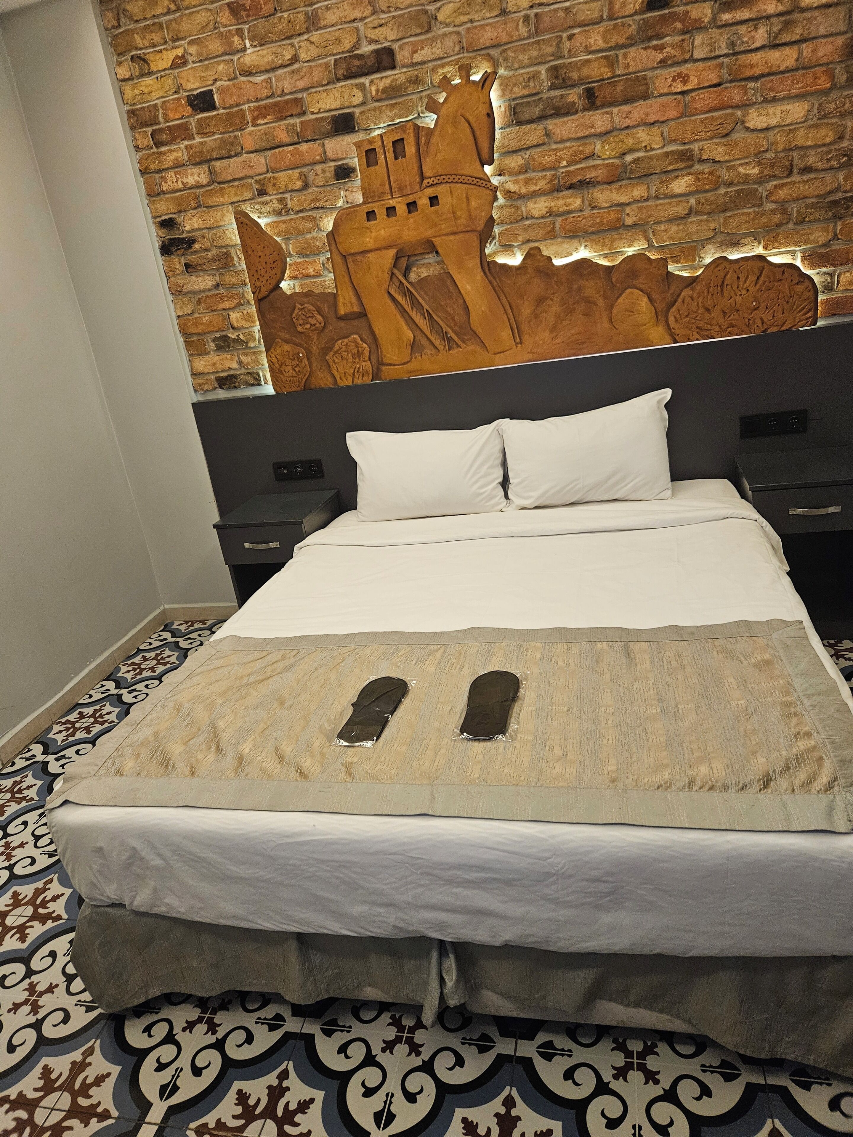 A hotel room with a brick wall, a bed with white bedding, a patterned floor, and a headboard with a horse and castle design.