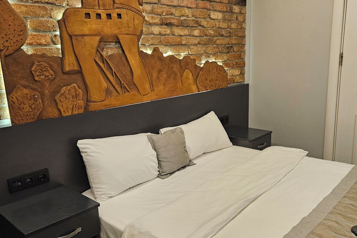 A hotel room with a brick wall, a bed with white linens, a nightstand, and a patterned floor.