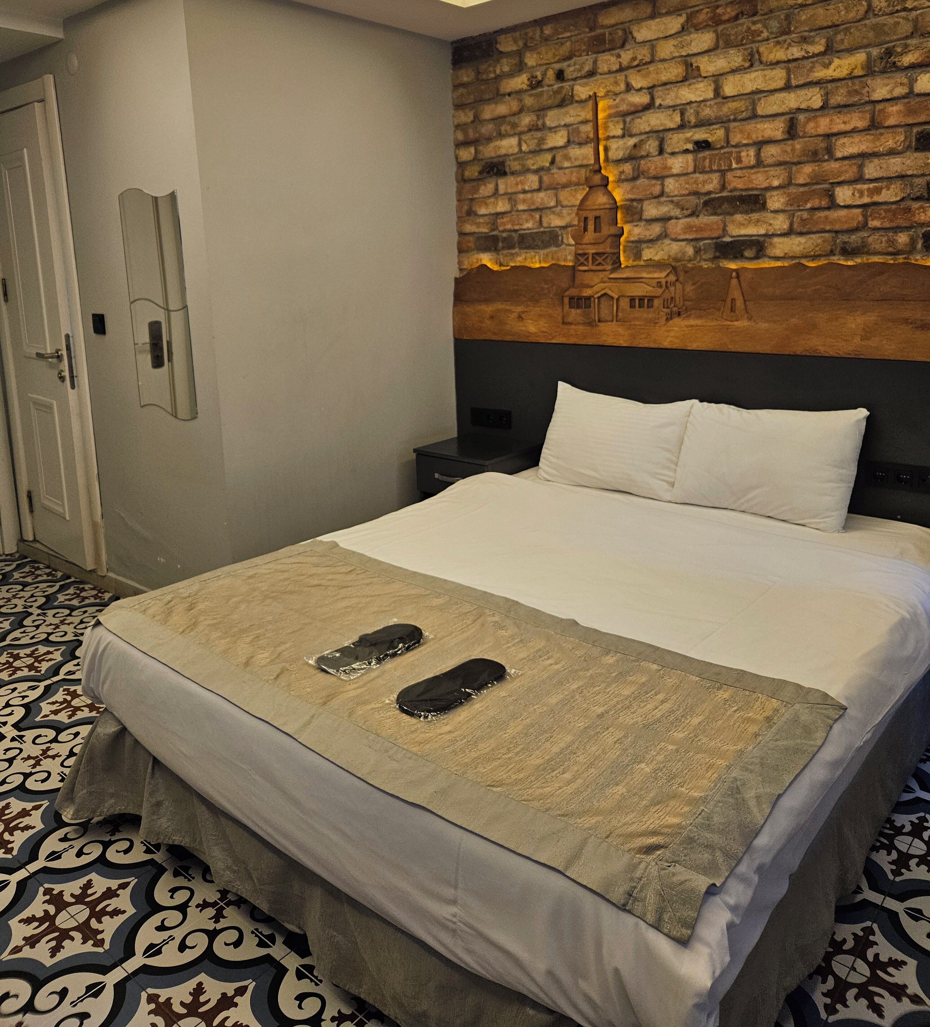 A hotel room with a bed, a brick wall mural, a patterned floor, and a door.