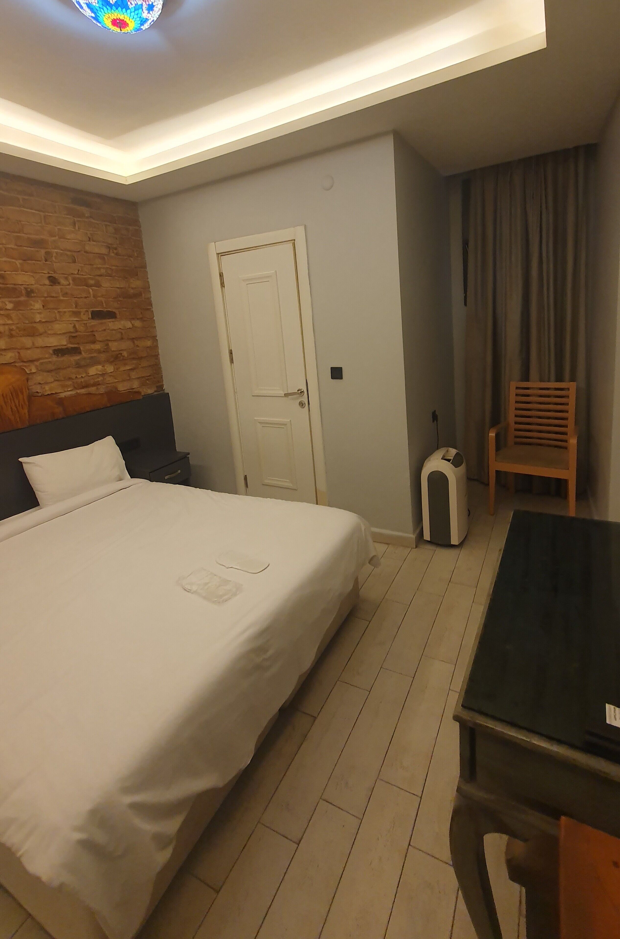 A hotel room with a large bed, a wooden headboard, a small bedside table, a desk, a chair, a suitcase, and a door.