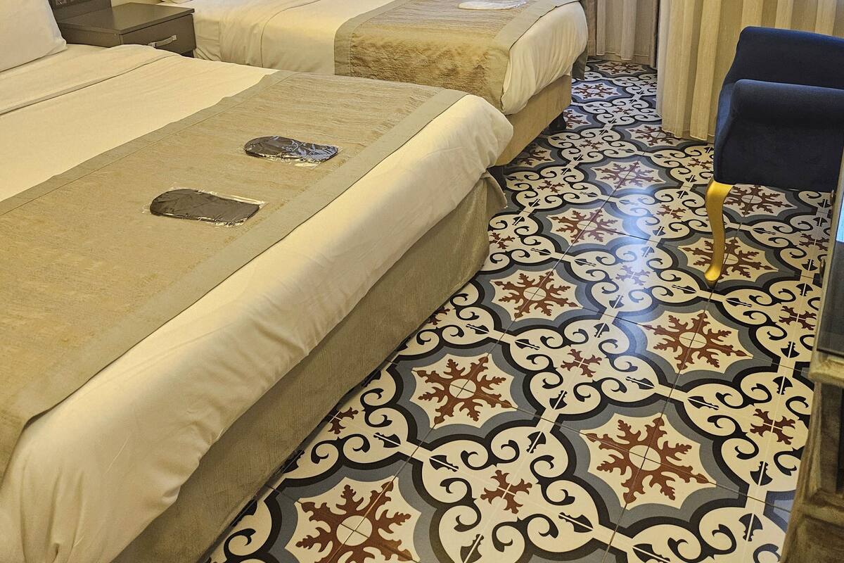 A hotel room with two beds, a desk, a chair, and patterned floor tiles.