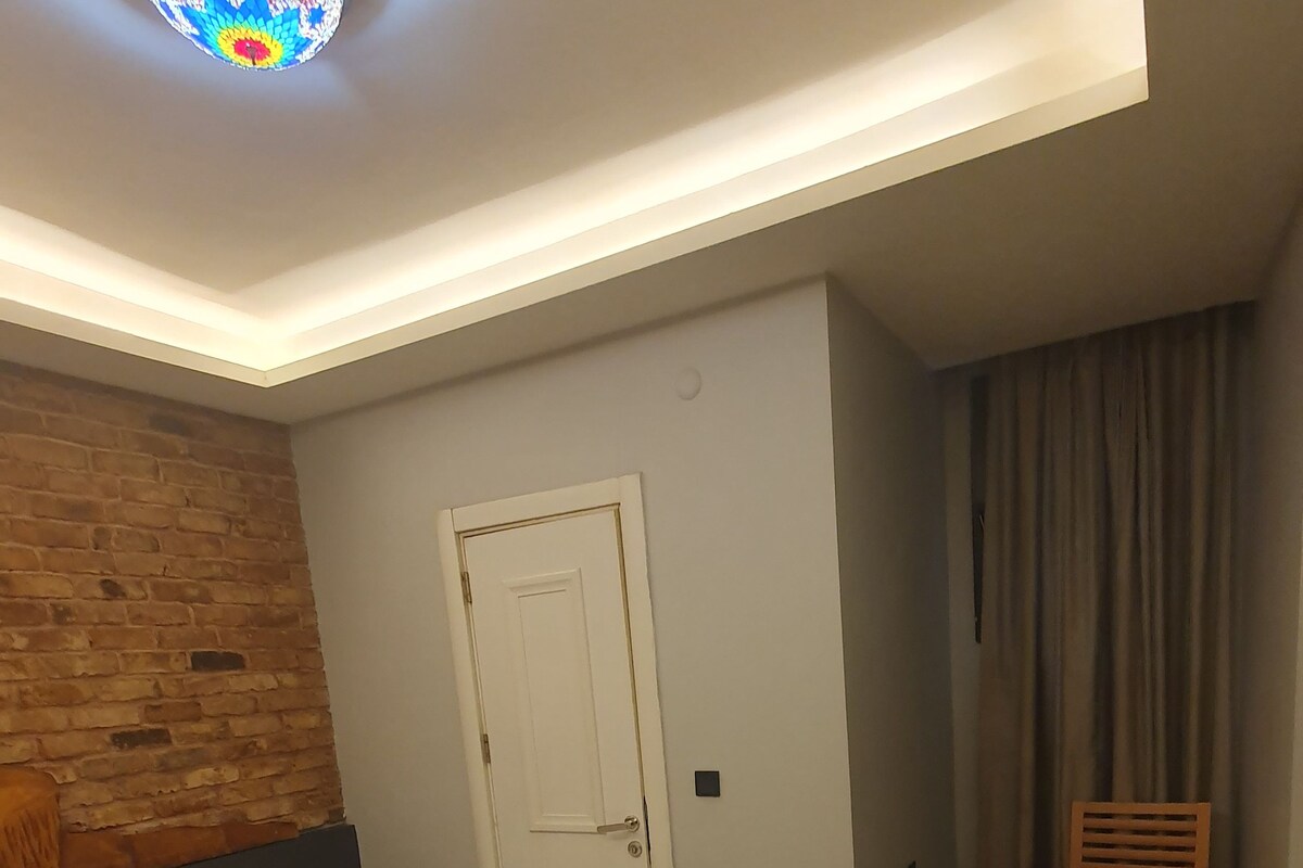 A hotel room with a large bed, a wooden headboard, a small bedside table, a desk, a chair, a suitcase, and a door.