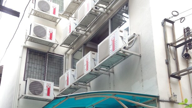 A building with multiple air conditioning units mounted on the exterior wall, a Coca-Cola refrigerator, and a blue awning over a doorway.