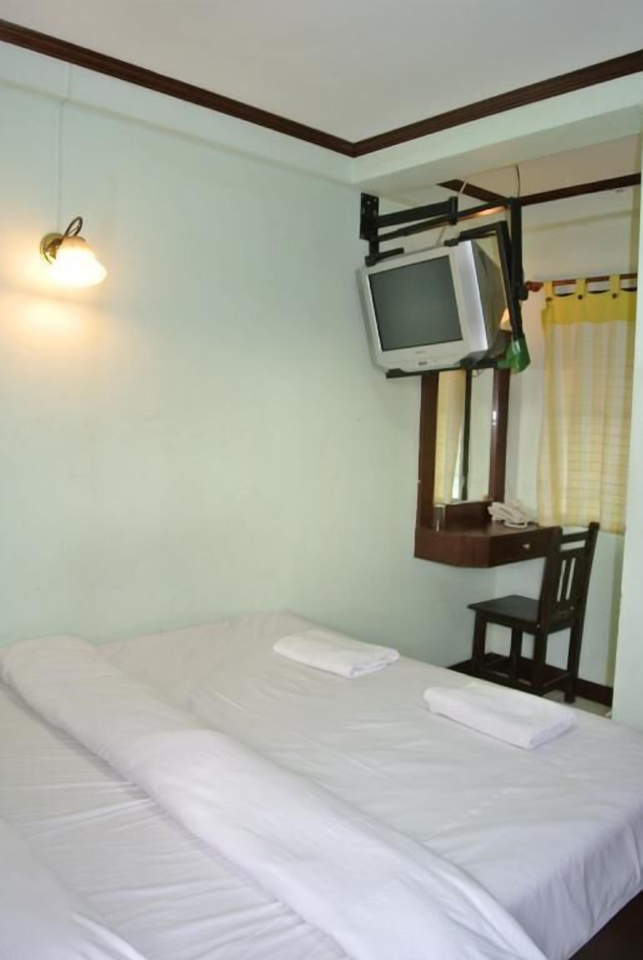 A single bed with white linens, a desk with a mirror, a television mounted above it, and a wall-mounted light fixture.