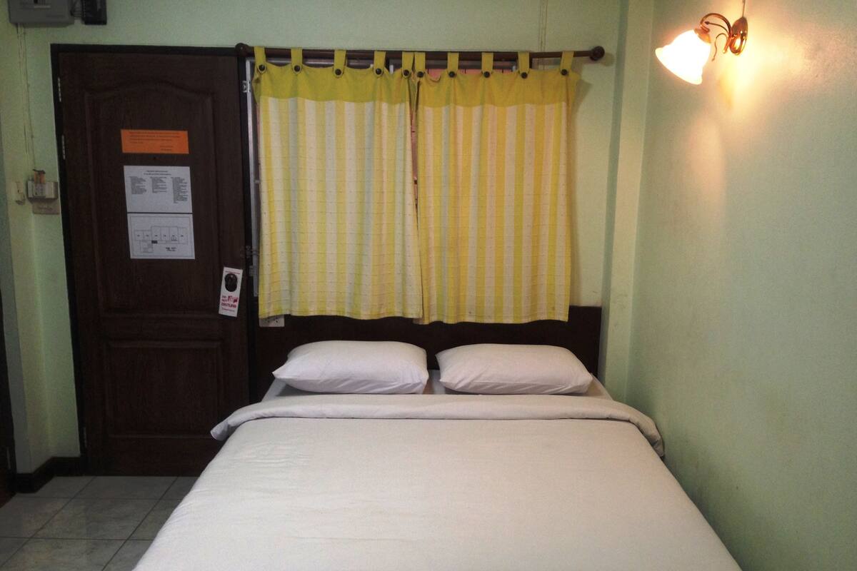 Um quarto simples com uma cama, uma porta com uma placa e uma janela com cortinas amarelas.