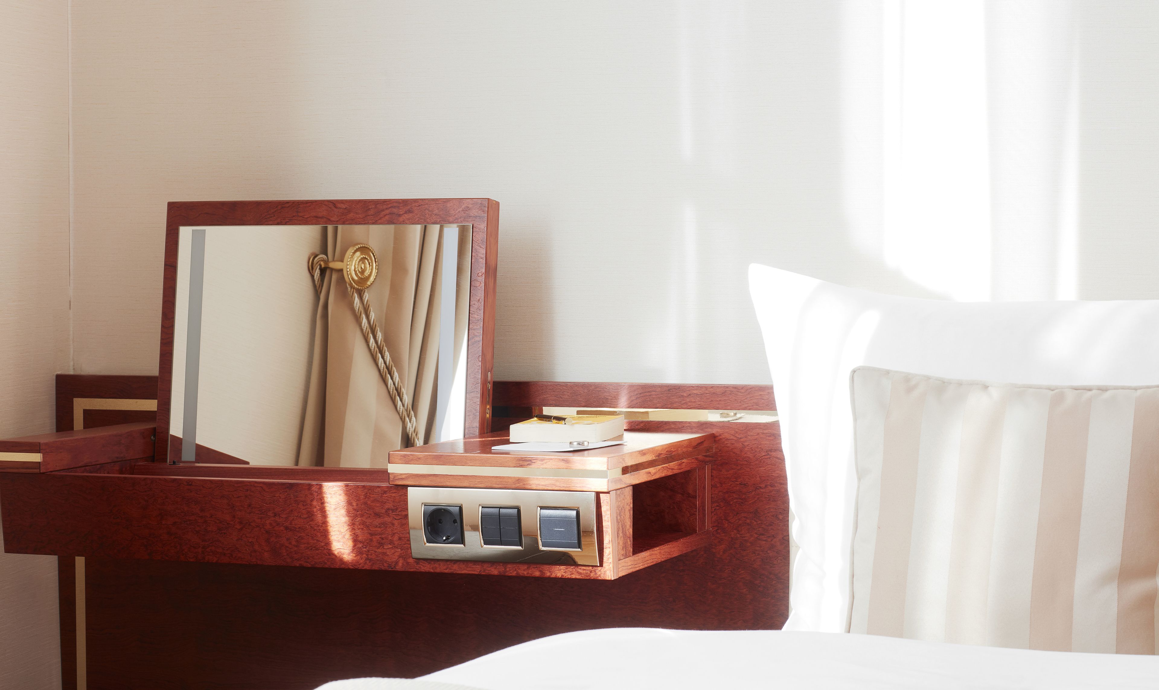 A hotel room with a wooden nightstand, a mirror, a bedside table, and a bed with white bedding.