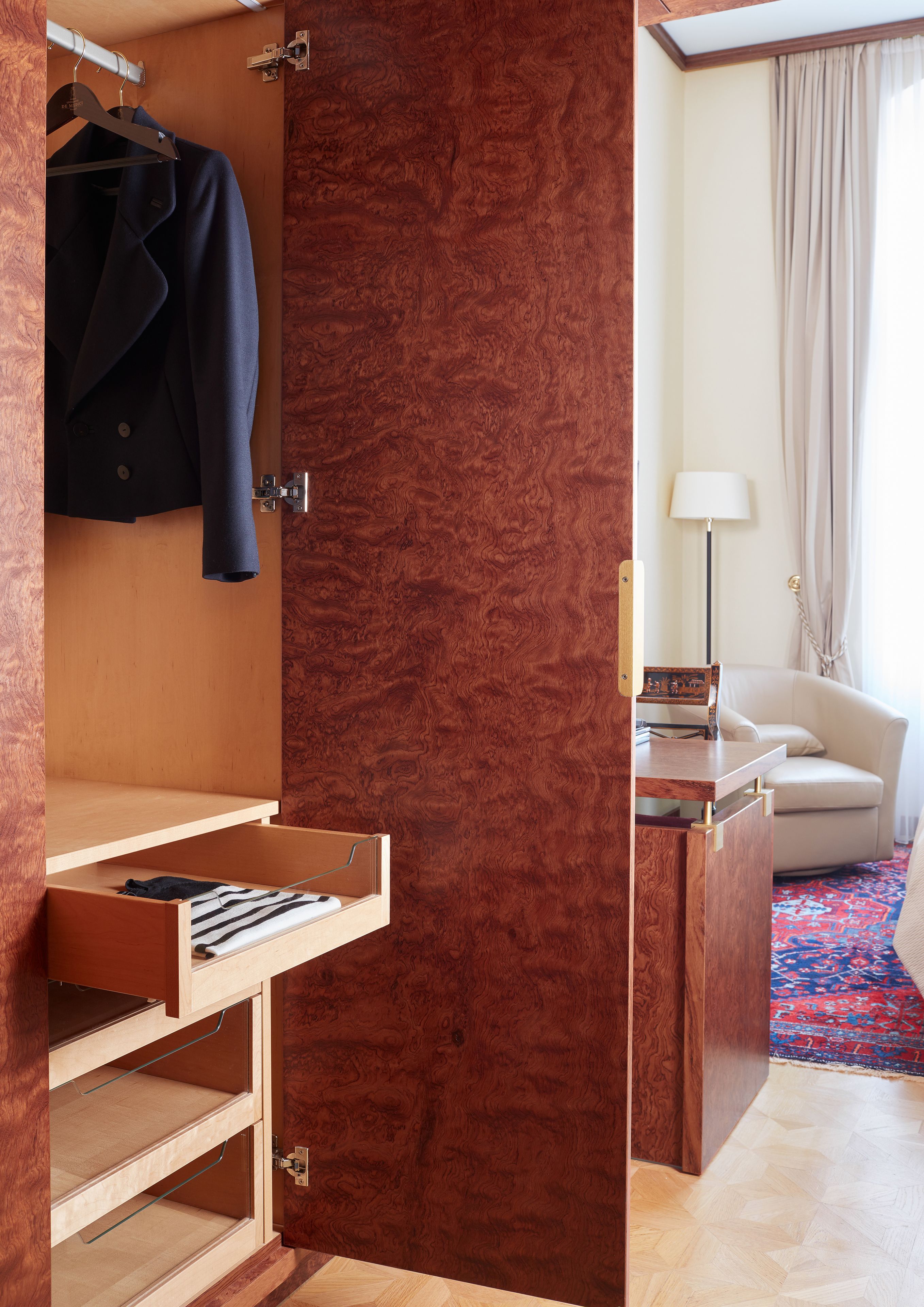 A hotel room with a wooden wardrobe, a desk with a lamp, a sofa, and a patterned carpet.