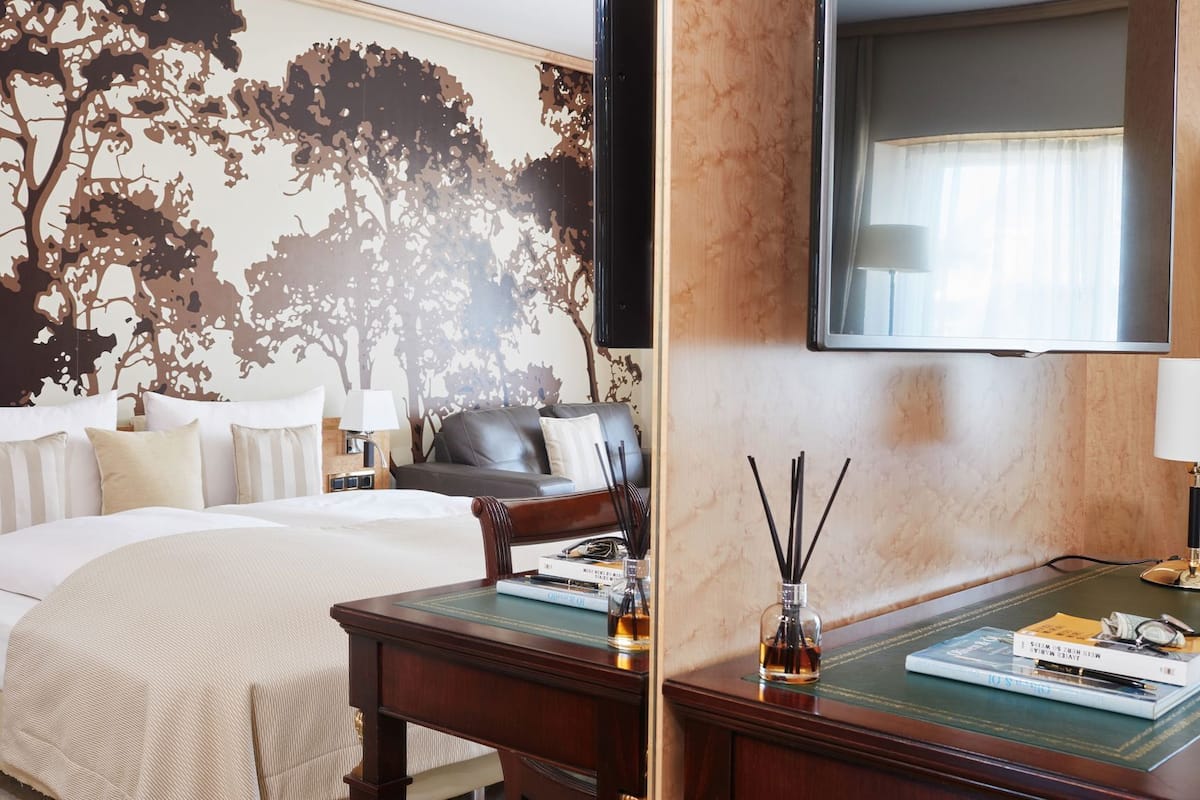 Una habitación de hotel con cama, escritorio, silla, televisor y una pared con un mural de un árbol.