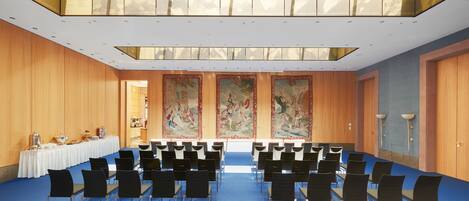 A conference room with rows of chairs, a long table with a white tablecloth, and a large tapestry on the wall.