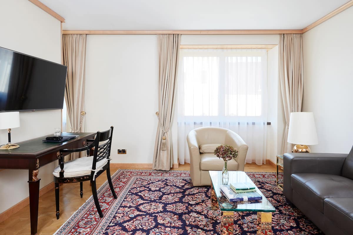 Habitación de hotel con televisión, escritorio, silla, sofá, mesa de centro y ventana grande con cortinas.