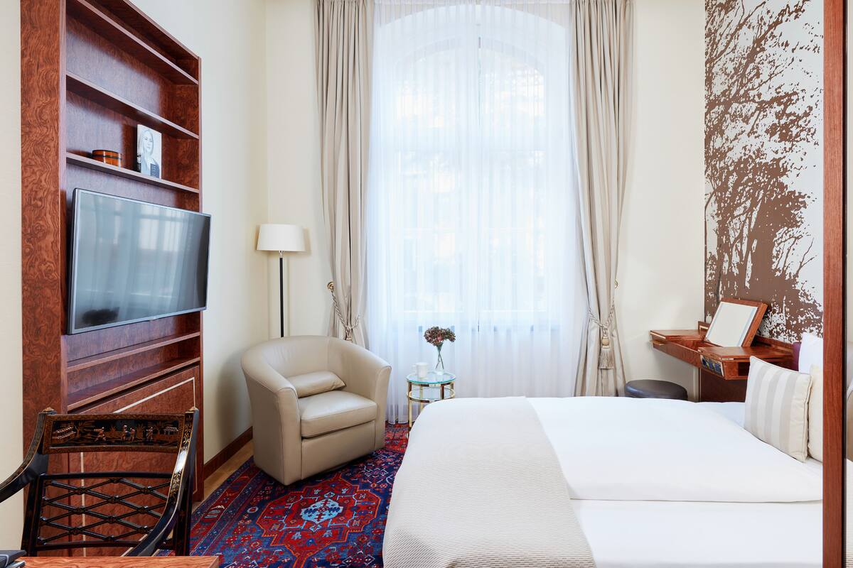 Habitación de hotel con cama, televisión, una silla, una mesita con un florero y un ventanal con cortinas.