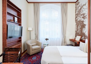 Ein Hotelzimmer mit Bett, Fernseher, Sessel, kleinem Tisch mit Vase und großem Fenster mit Vorhängen.