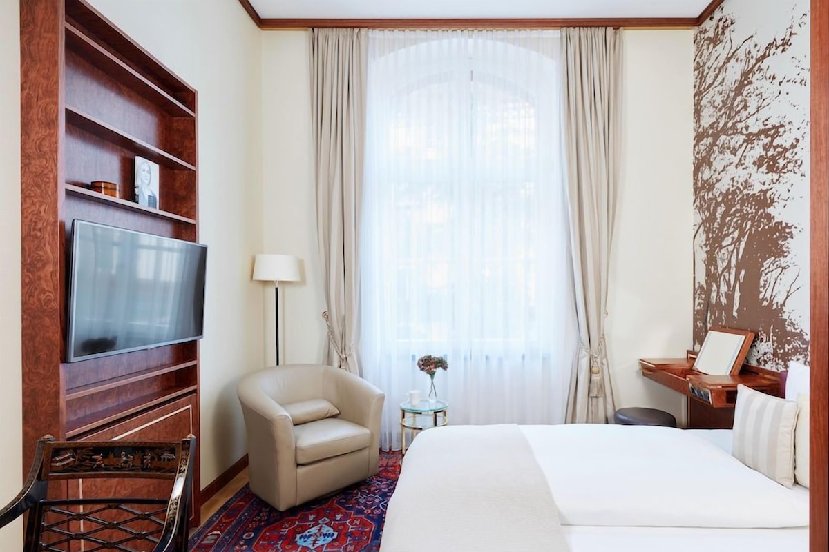 Una habitación de hotel con una cama, televisión, sillón, escritorio y ventana con cortinas.