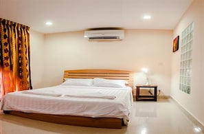 Una cama bien tendida en una habitación de hotel con cabecera de madera, unidad de aire acondicionado montada en la pared, mesita de noche con lámpara y ventana con persianas.