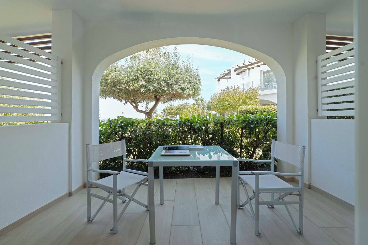 A patio with a white table and chairs, an archway, and a view of greenery and a building in the background.