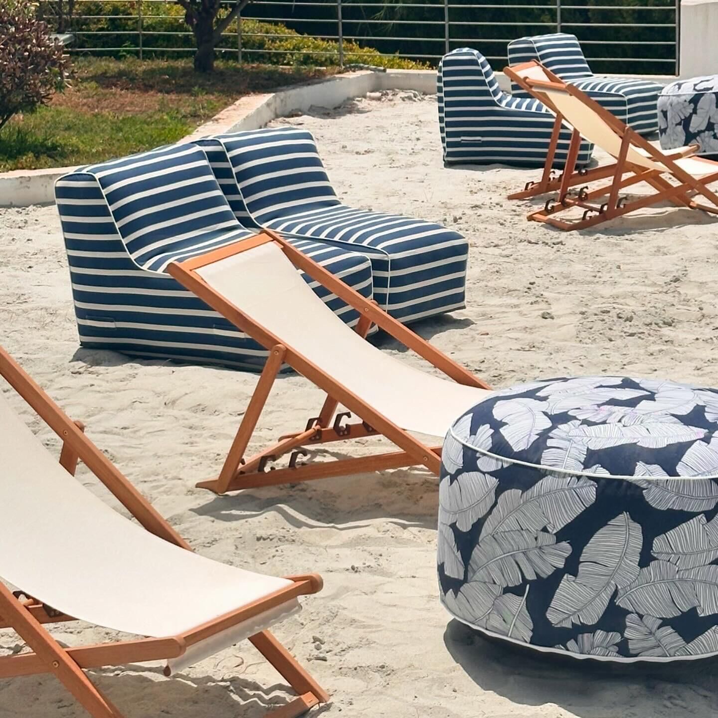A beachside lounge area with striped lounge chairs and ottomans, arranged on a sandy surface.