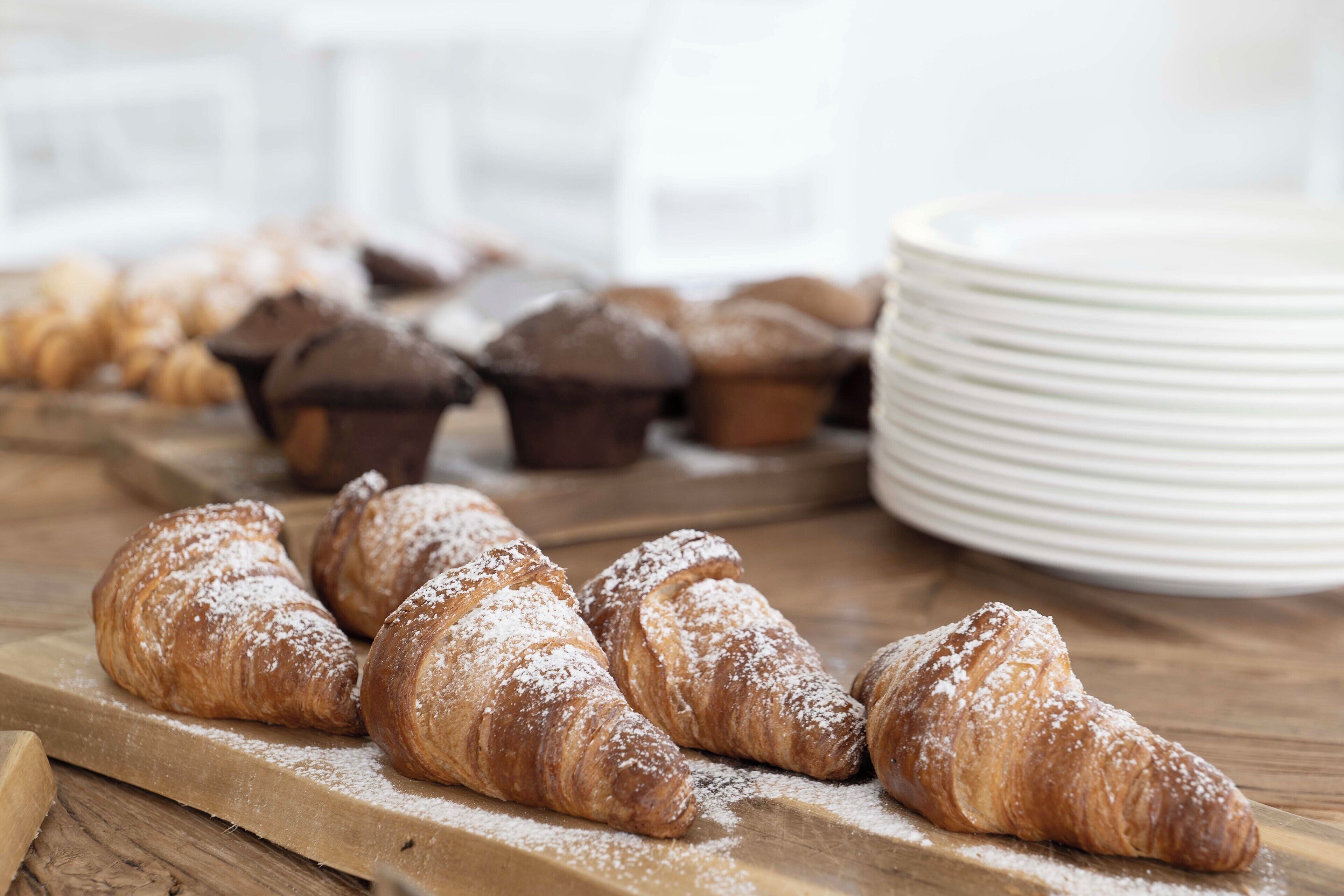 Croissants and muffins on a wooden board with a stack of white plates in the background.