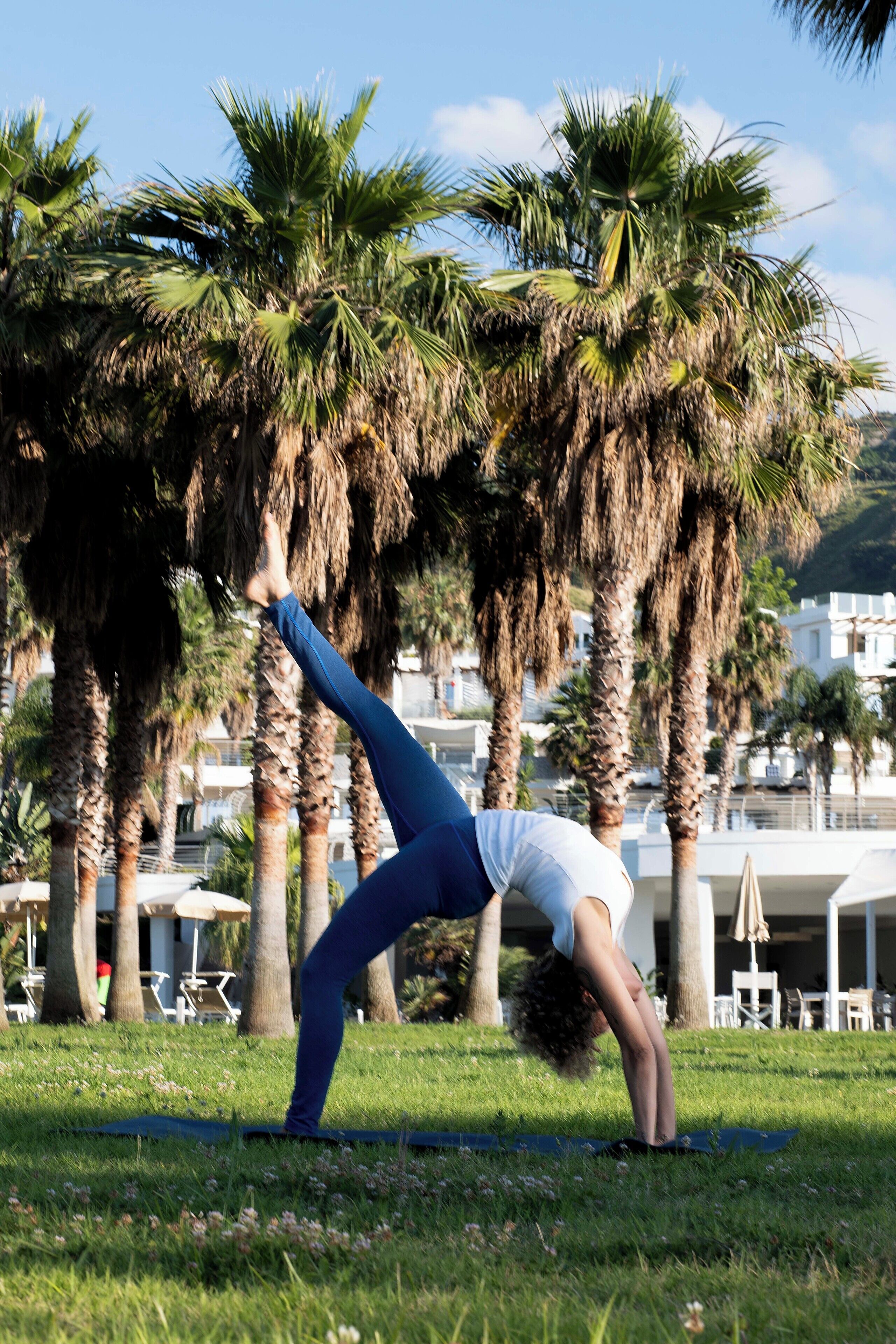 Eine Person führt eine Yoga-Pose auf einer Matte in einer grünen Umgebung mit Palmen und Gebäuden im Hintergrund aus.