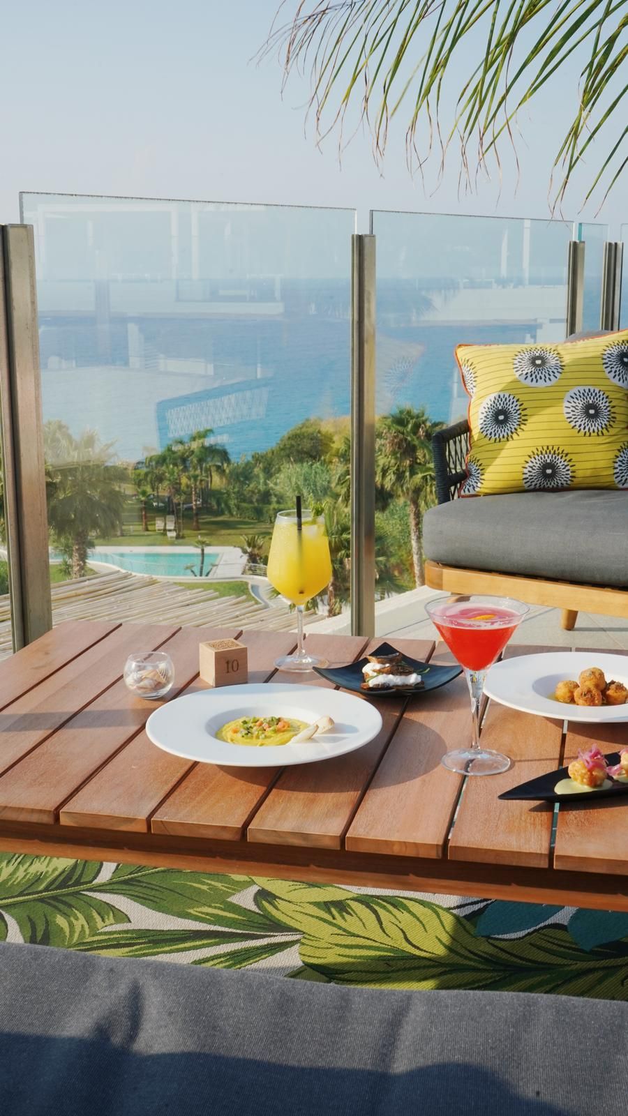 A wooden table with a view of a pool and palm trees, a yellow pillow, and a martini glass.