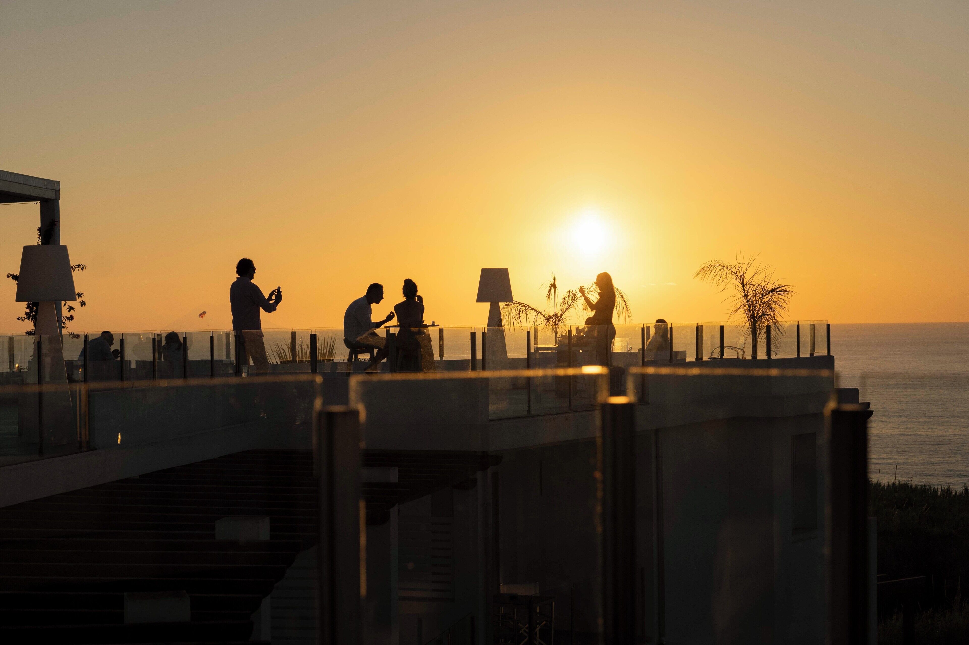 Vista al atardecer desde un balcón con vista al mar, donde gente conversa y participa en diversas actividades.