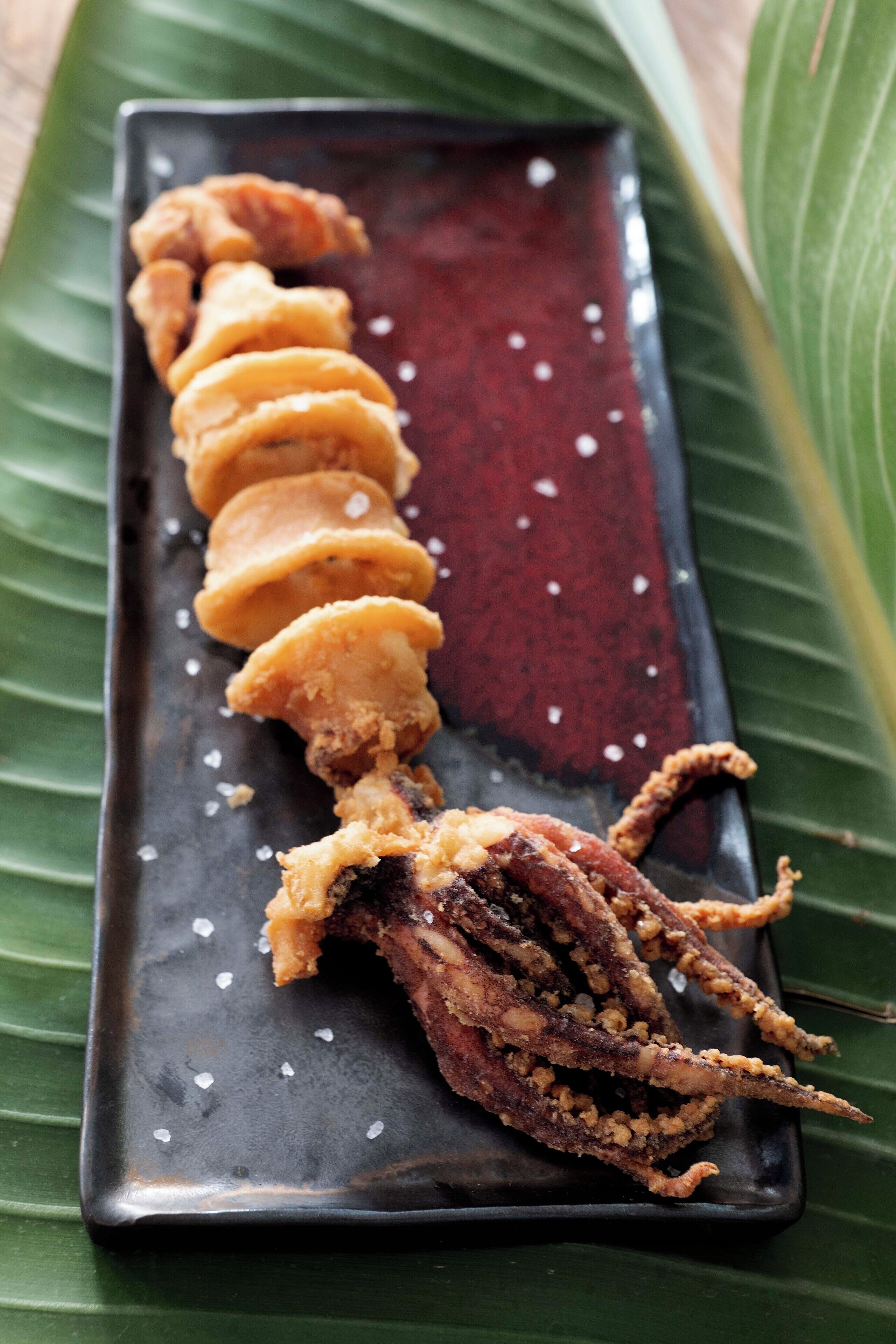 A rectangular plate with a row of fried seafood items, garnished with salt, on a dark surface with a green leaf underneath.