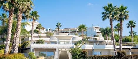 Un edificio residencial de varios pisos con un estilo arquitectónico moderno, con balcones y un jardín bien cuidado con palmeras.
