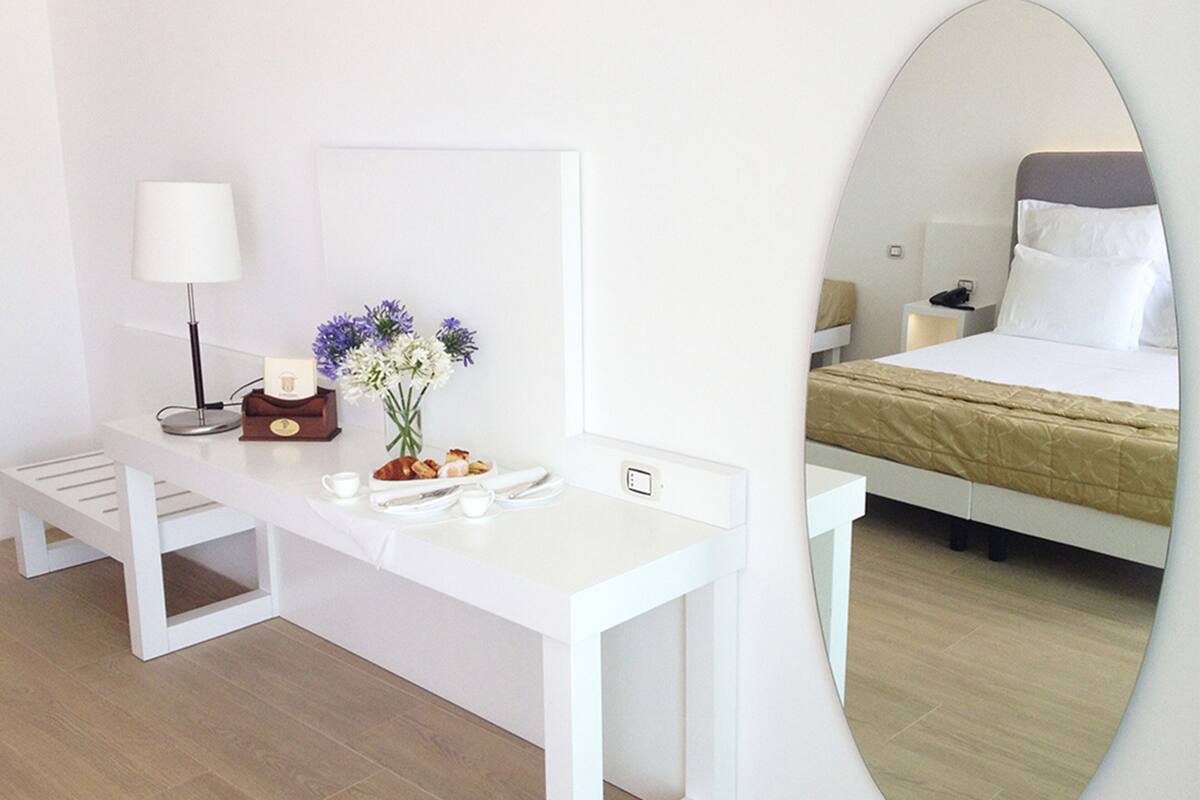 A hotel room with a white desk, a lamp, a vase with flowers, and a bed with a headboard.