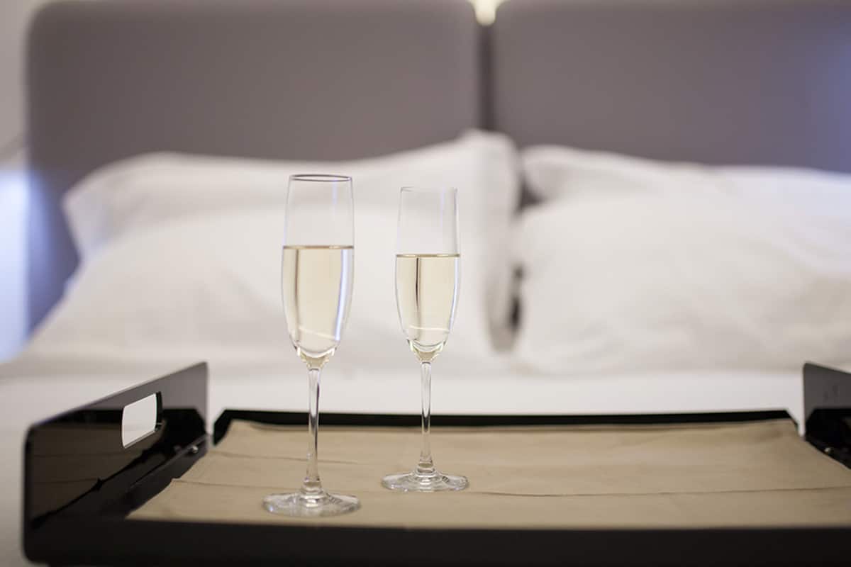 Two champagne flutes on a tray in front of a bed with white linens and a gray headboard.
