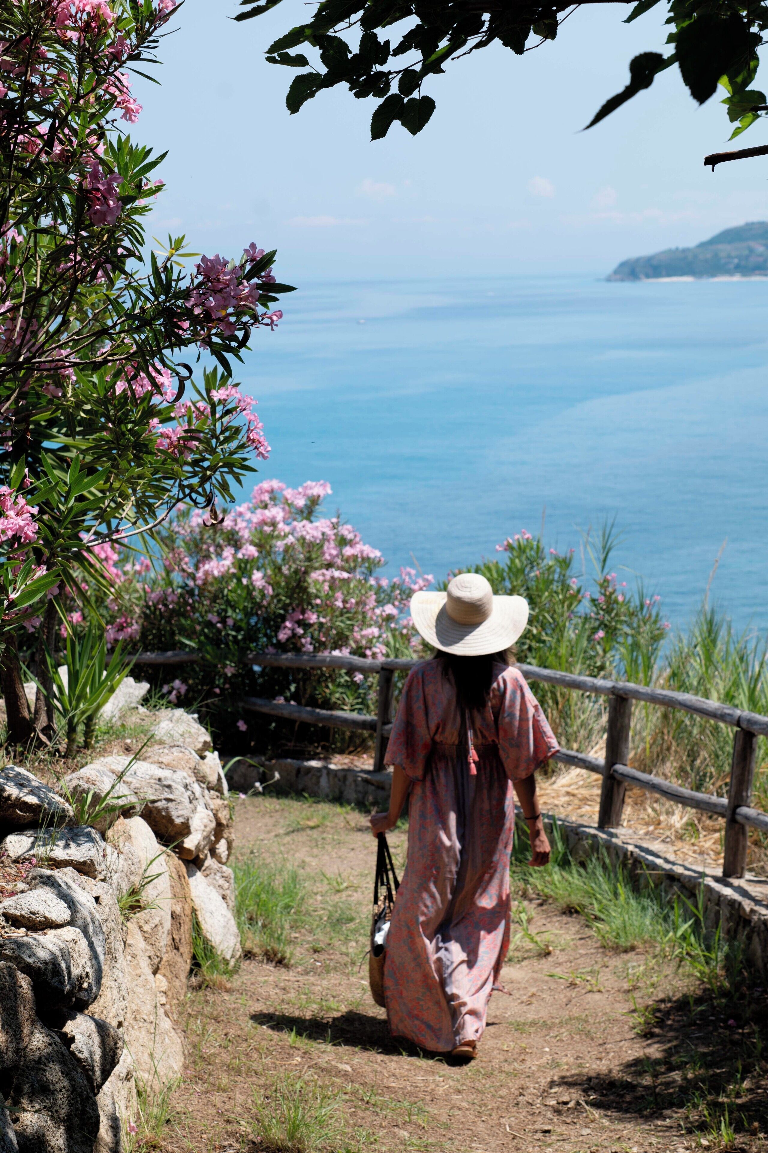 A woman in a long dress and hat walking on a dirt path beside a wooden fence, overlooking a coastal view.