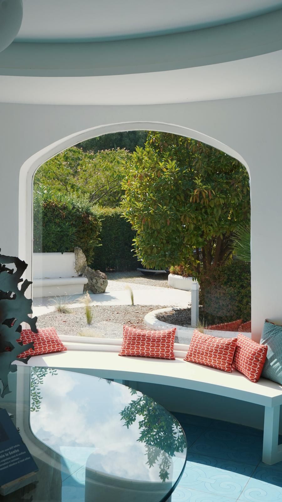 Een moderne kamer met een groot rondboogvenster met uitzicht op een tuin. Op de voorgrond staat een witte bank met rode, gemêleerde kussens en een glazen tafel.