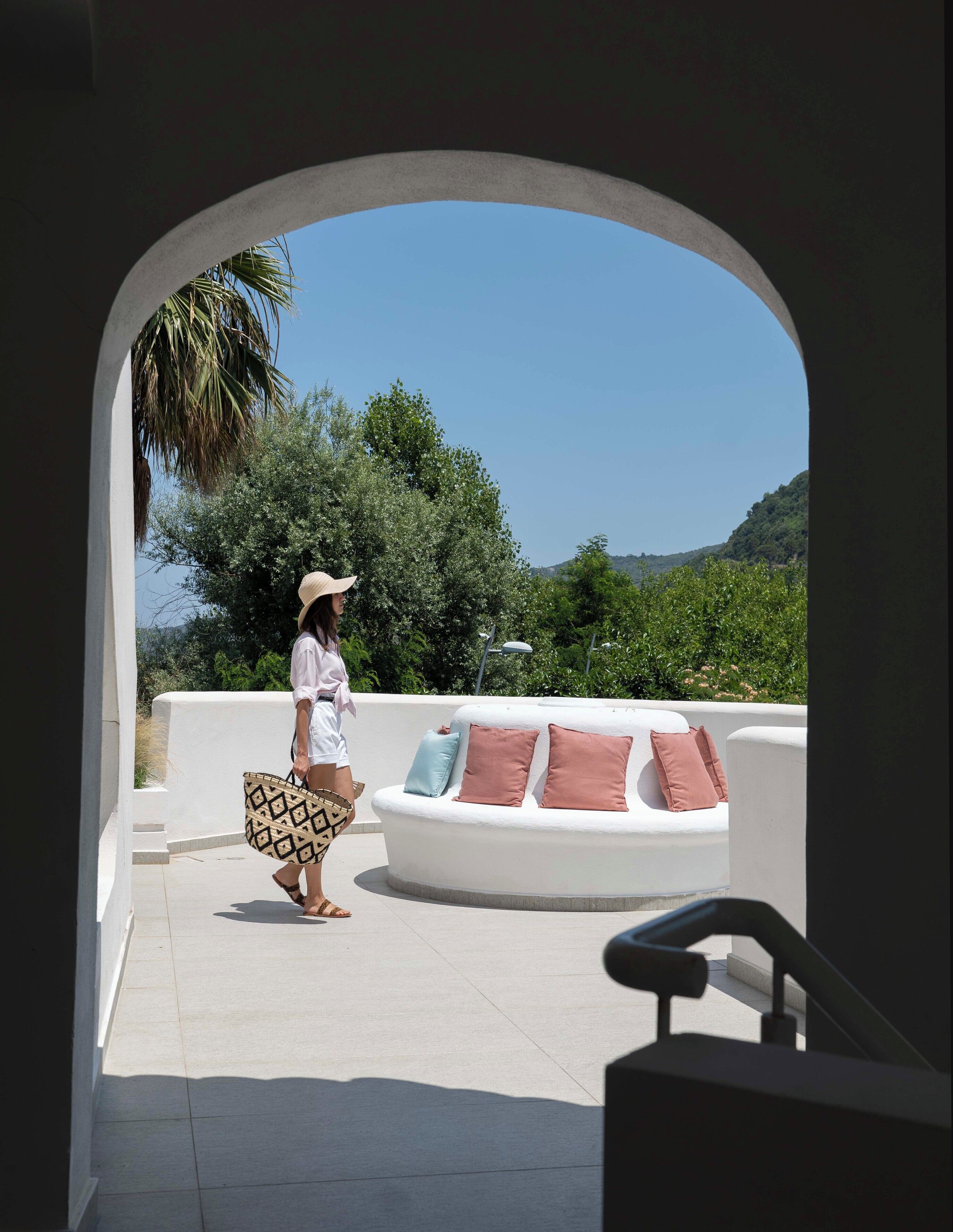 A person walking through a white archway onto a terrace with a white sofa and cushions, overlooking a green landscape.