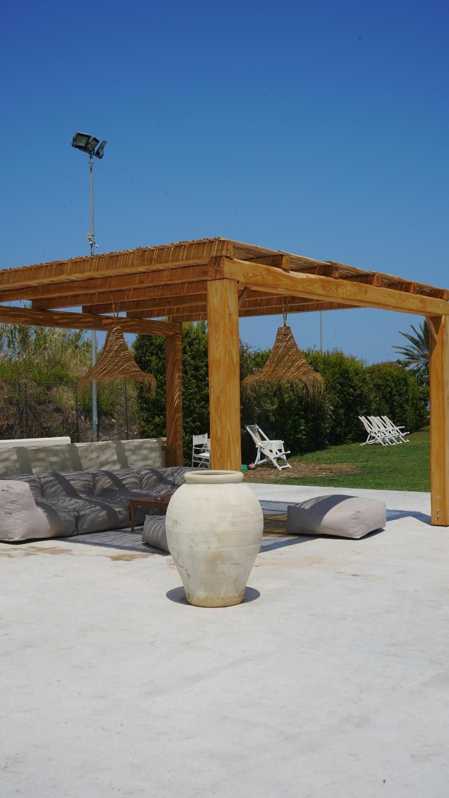 Une structure en bois formant une pergola, avec un grand vase blanc en premier plan, entourée de fauteuils et surmontée d’un toit de chaume.