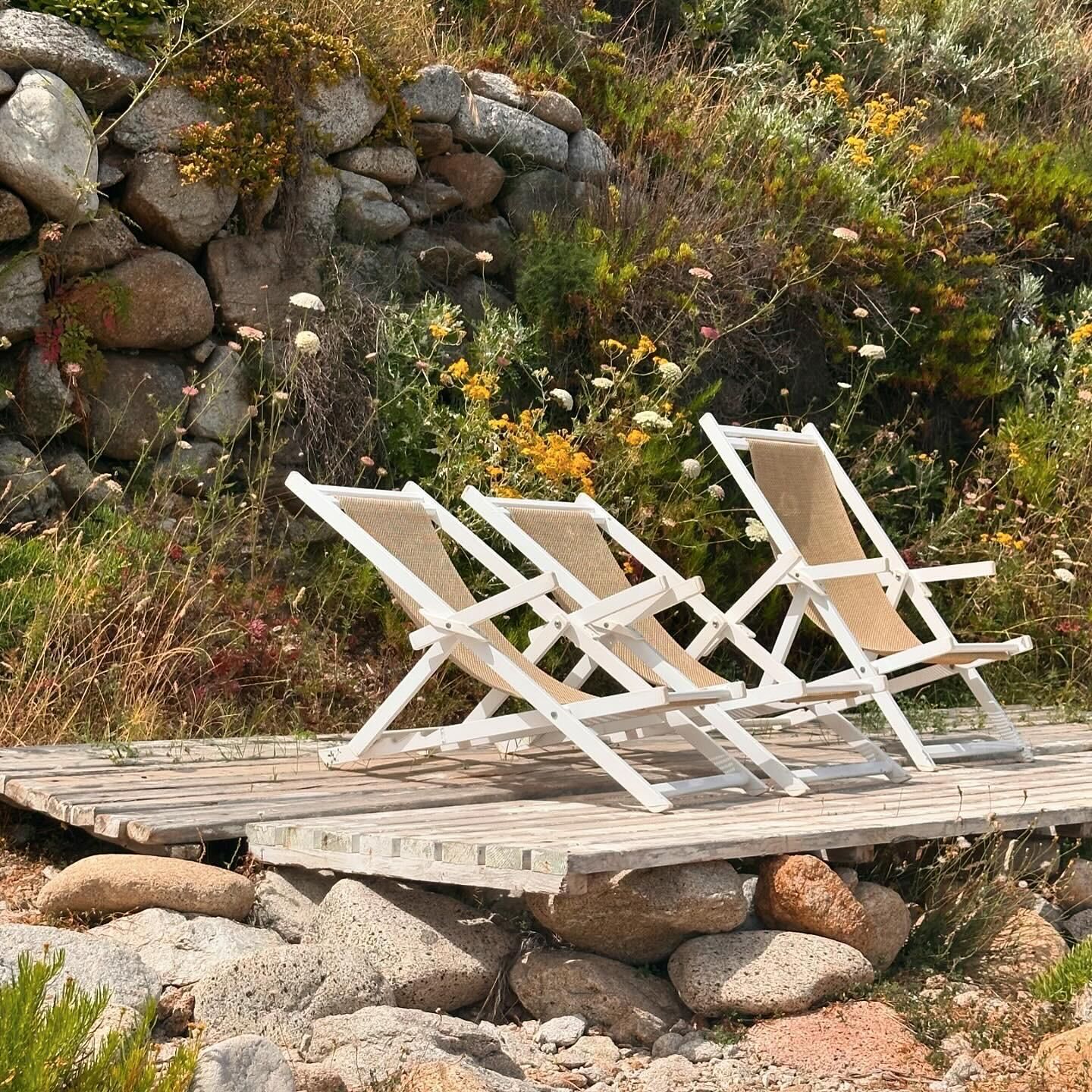 Tres sillas blancas de descanso sobre una terraza de madera, con un fondo rocoso y cubierto de vegetación.