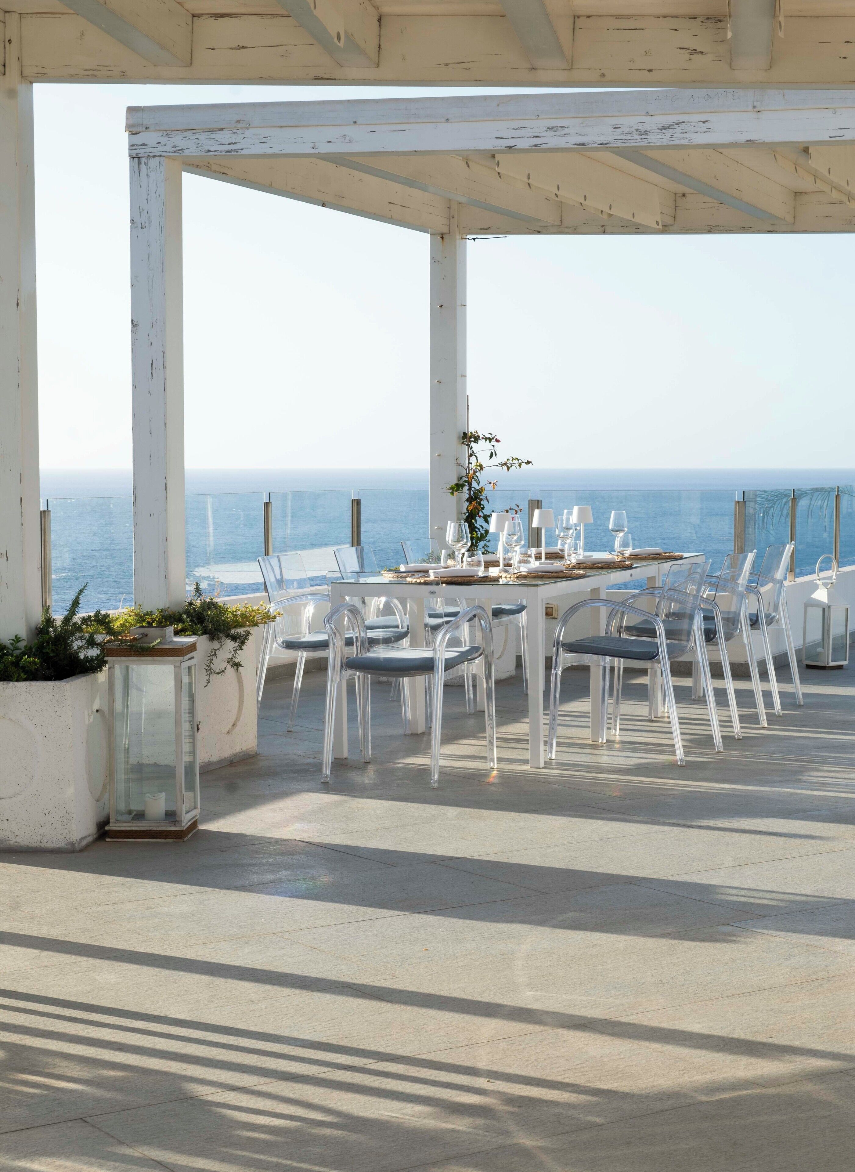 Un área de comedor al aire libre con una mesa dispuesta para seis personas, sillas transparentes y vistas al mar.