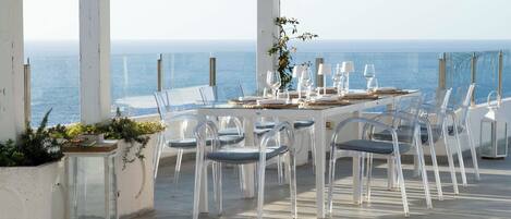 Un área de comedor al aire libre con una mesa dispuesta para seis personas, sillas transparentes y vistas al mar.