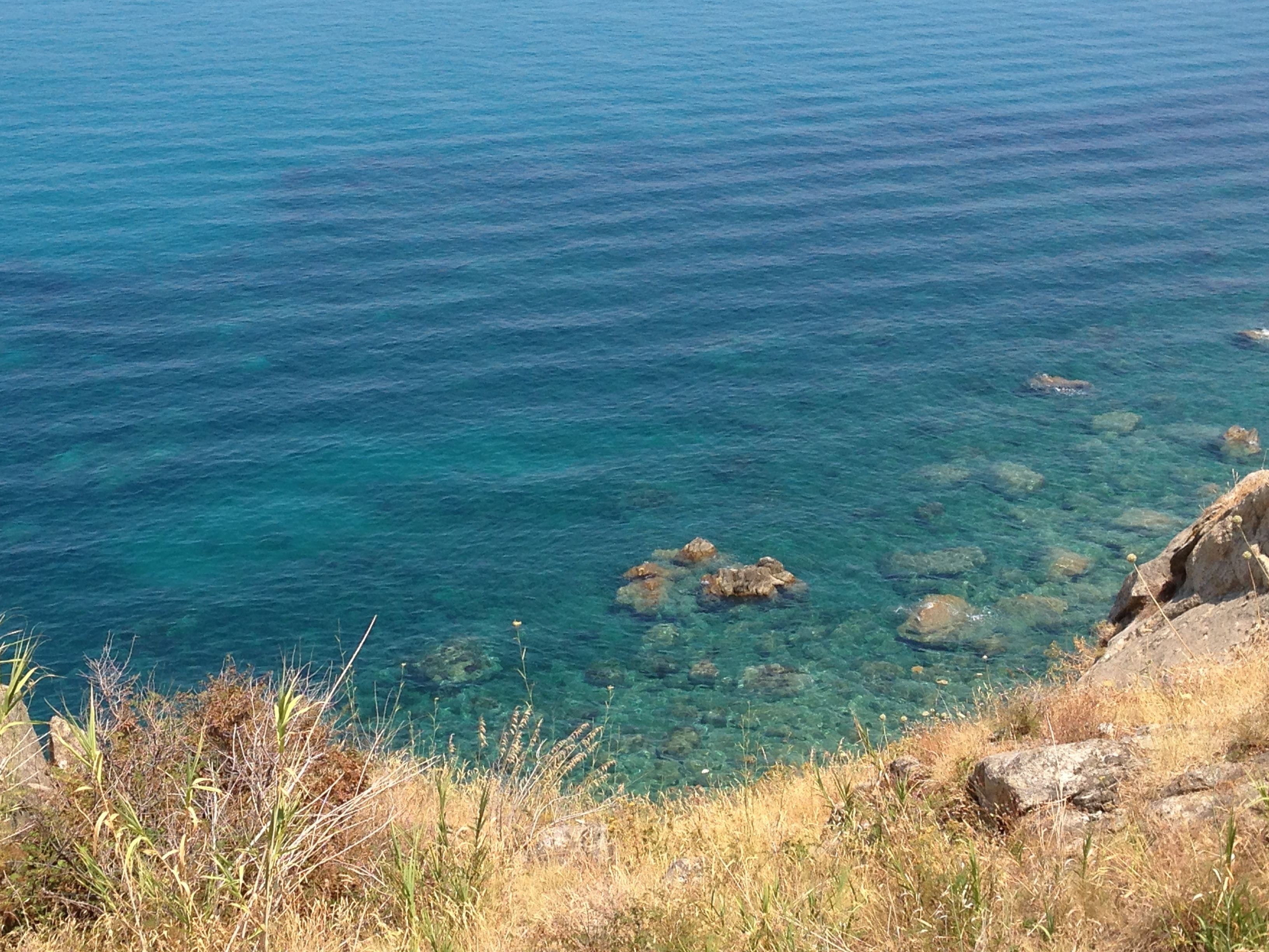 A coastal landscape with clear blue water, rocky outcrops, and dry grass.