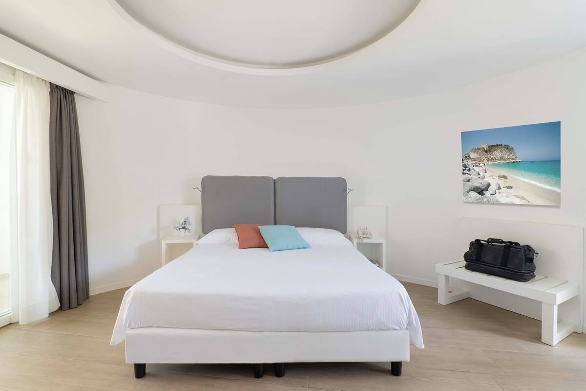 A modern hotel room with a large bed, bedside tables, a bench, and a painting of a beach.