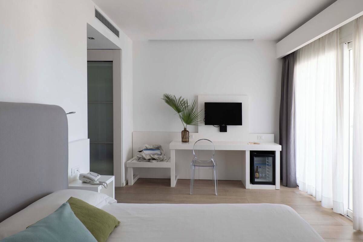A modern hotel room with a bed, a desk, a chair, a television, and a small refrigerator.