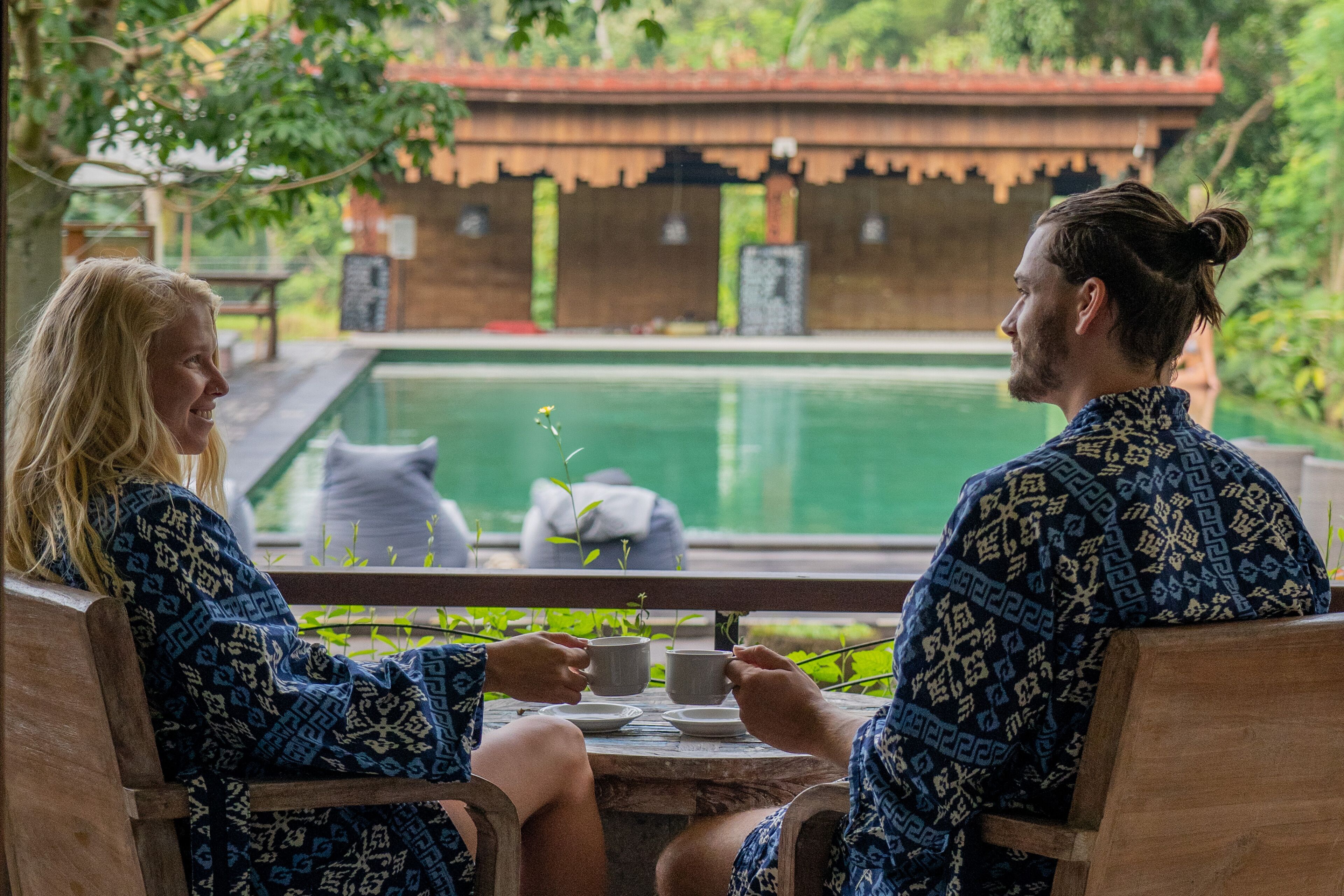 Two people sitting at a wooden table with a view of a swimming pool and a traditional building in the background.